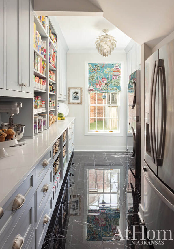 A bright pantry with pale blue cabinets, a long countertop, and a mixer sets the scene. Shelves filled with various canned goods line the left wall like headlines of culinary news. A window at the end features colorful curtains, next to a stainless steel fridge and glossy black floor tiles.